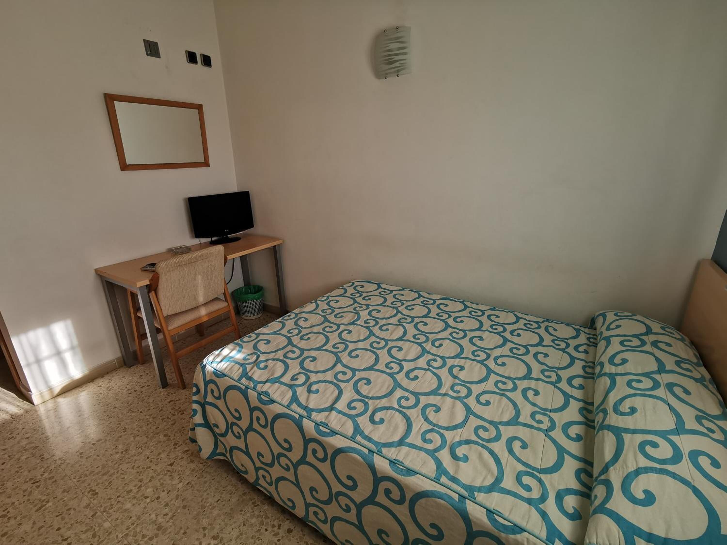 Un dormitorio con cama, escritorio, silla, televisor y espejo adosado a la pared.