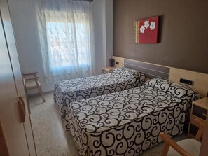 A bedroom with two beds, a bedside table, a chair, and a window with curtains. - Hostal Talavera (Talavera la Real)