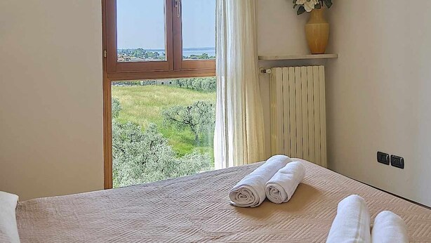 A hotel room with a large window, a bed with rolled towels, a radiator, and a vase with flowers on a shelf.
