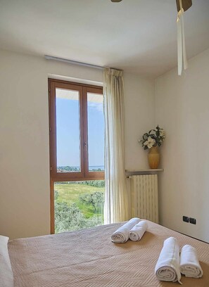A hotel room with a large window, a bed with rolled towels, a radiator, and a vase with flowers on a shelf.