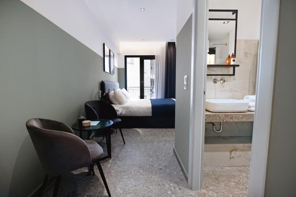 Habitación de hotel con cama, escritorio, silla, una mesita pequeña y baño con lavamanos y espejo.
