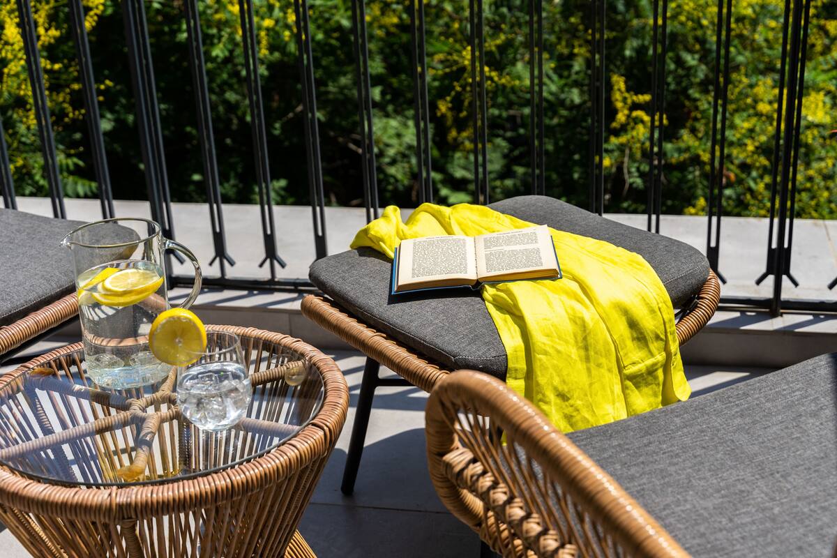A wicker chair with a cushion and a book, a glass table with a pitcher of water and a glass, and a yellow cloth on a lounge chair.