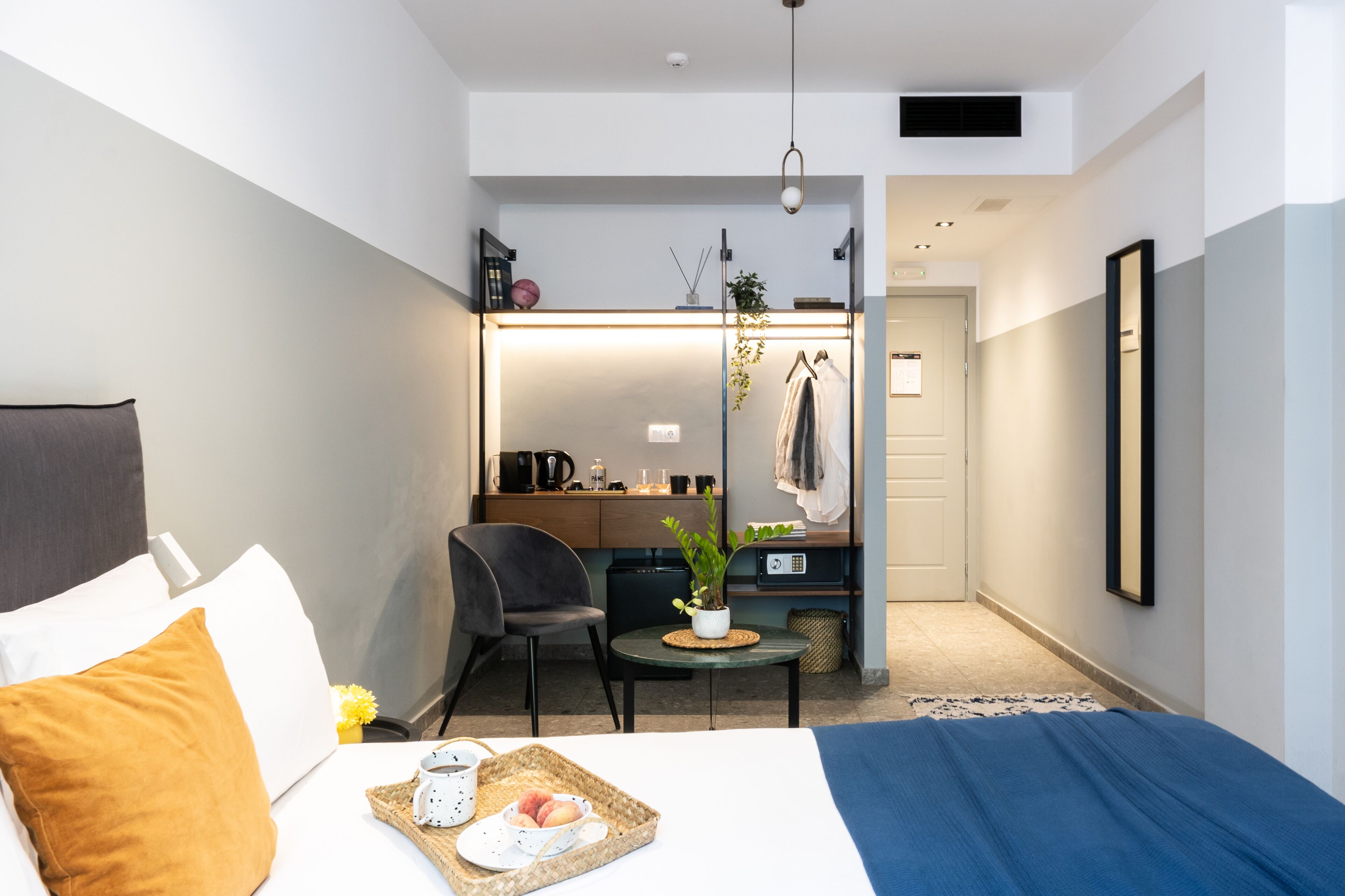 A hotel room with a bed, a desk, a chair, a small table with a tray of food, and a potted plant.