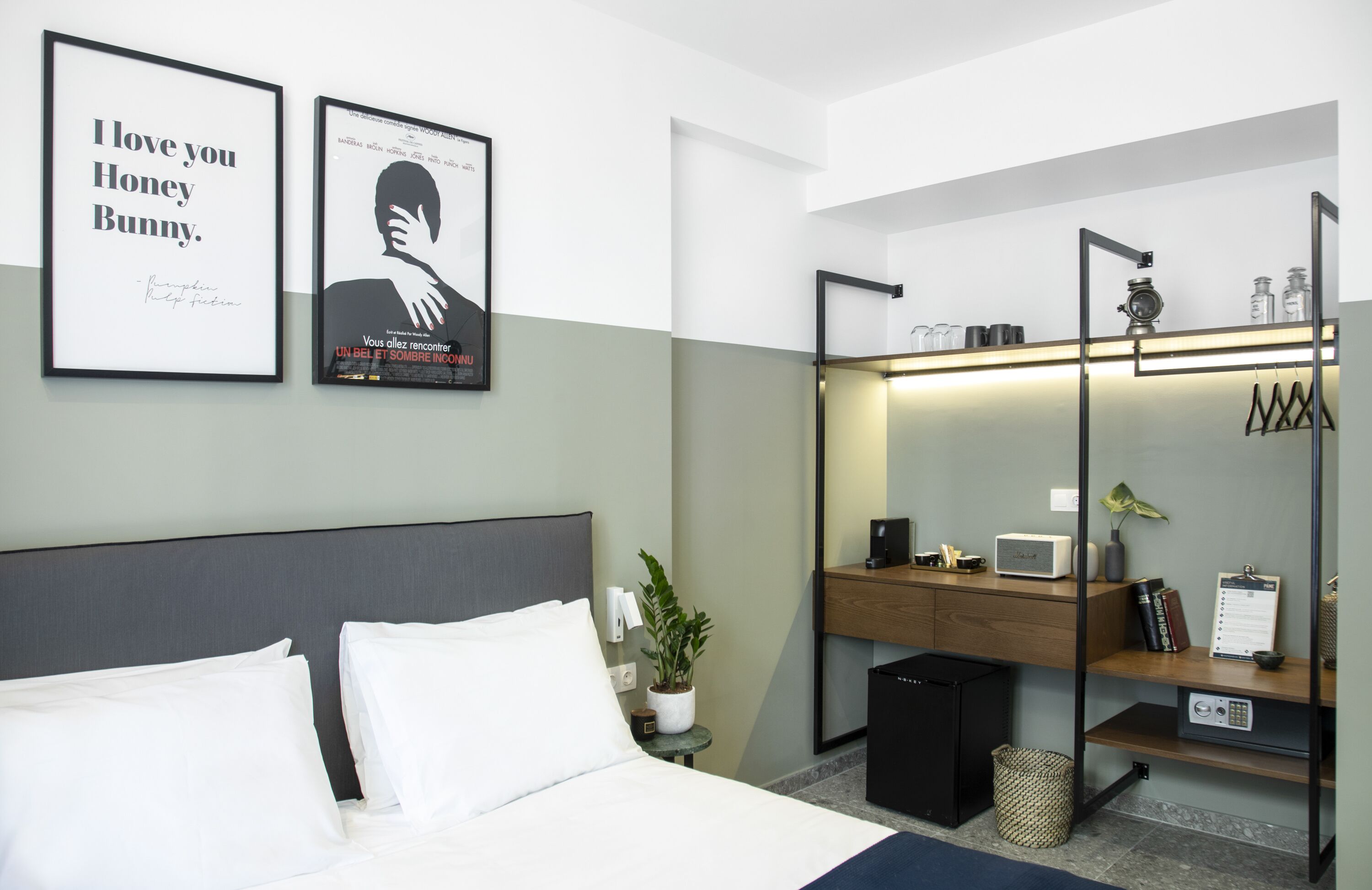A modern hotel room with a bed, bedside table, potted plant, and a built-in desk with a mirror and shelves.