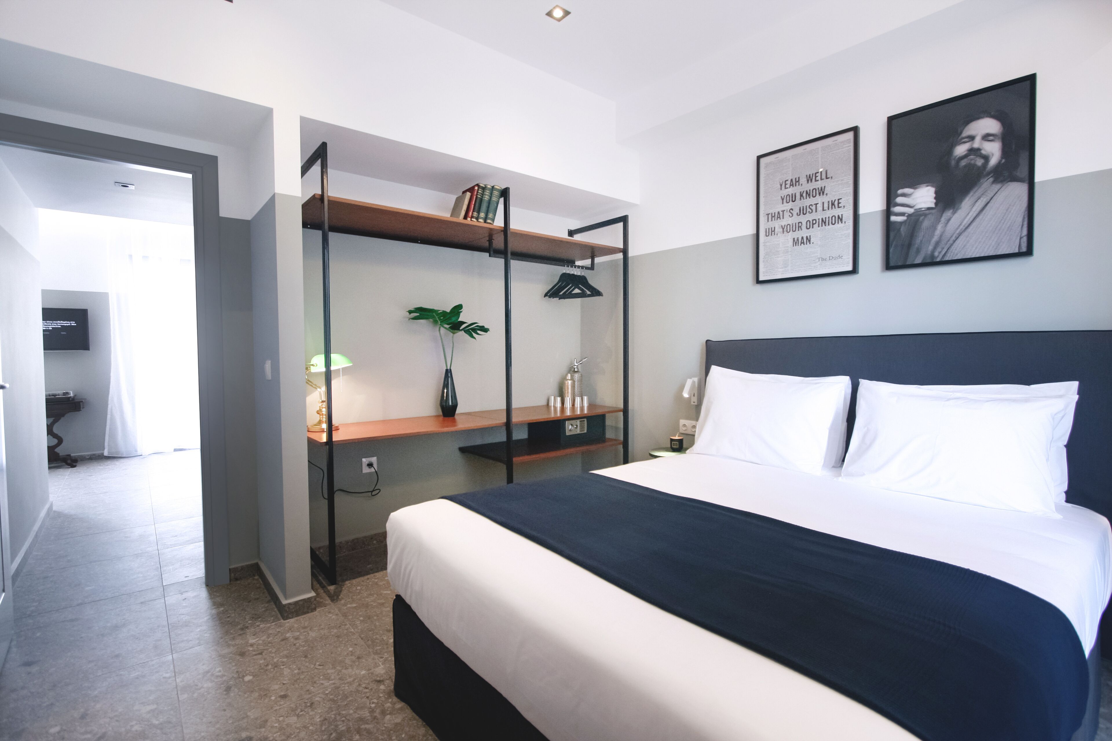 A modern hotel room with a large bed, a bedside table, a desk with a chair, a potted plant, and two framed quotes on the wall.