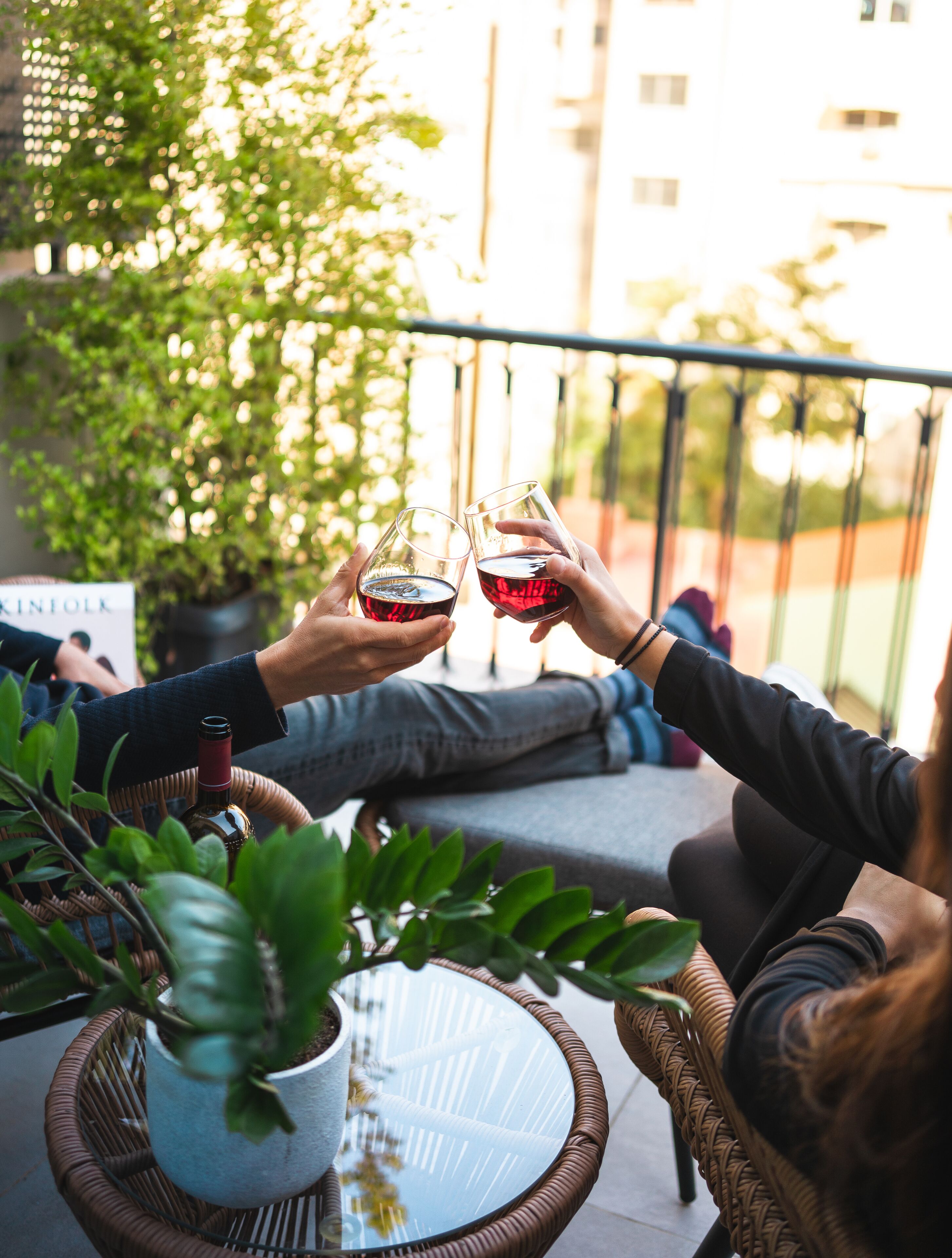 Twee personen proosten met wijn in een balkon met rieten meubels en plantenpotten.