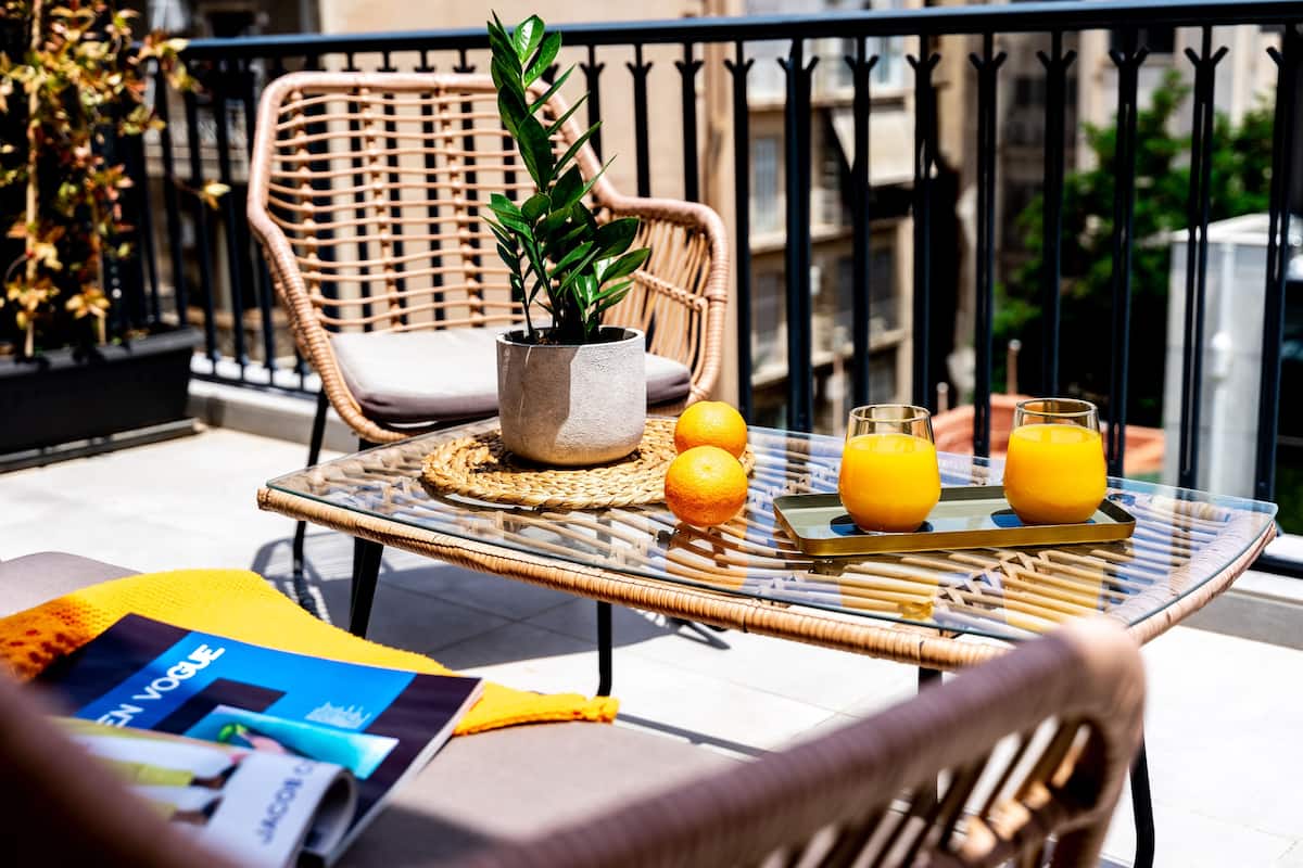 A wicker chair with a potted plant, a glass table with a tray of orange juice, and a magazine on a balcony.