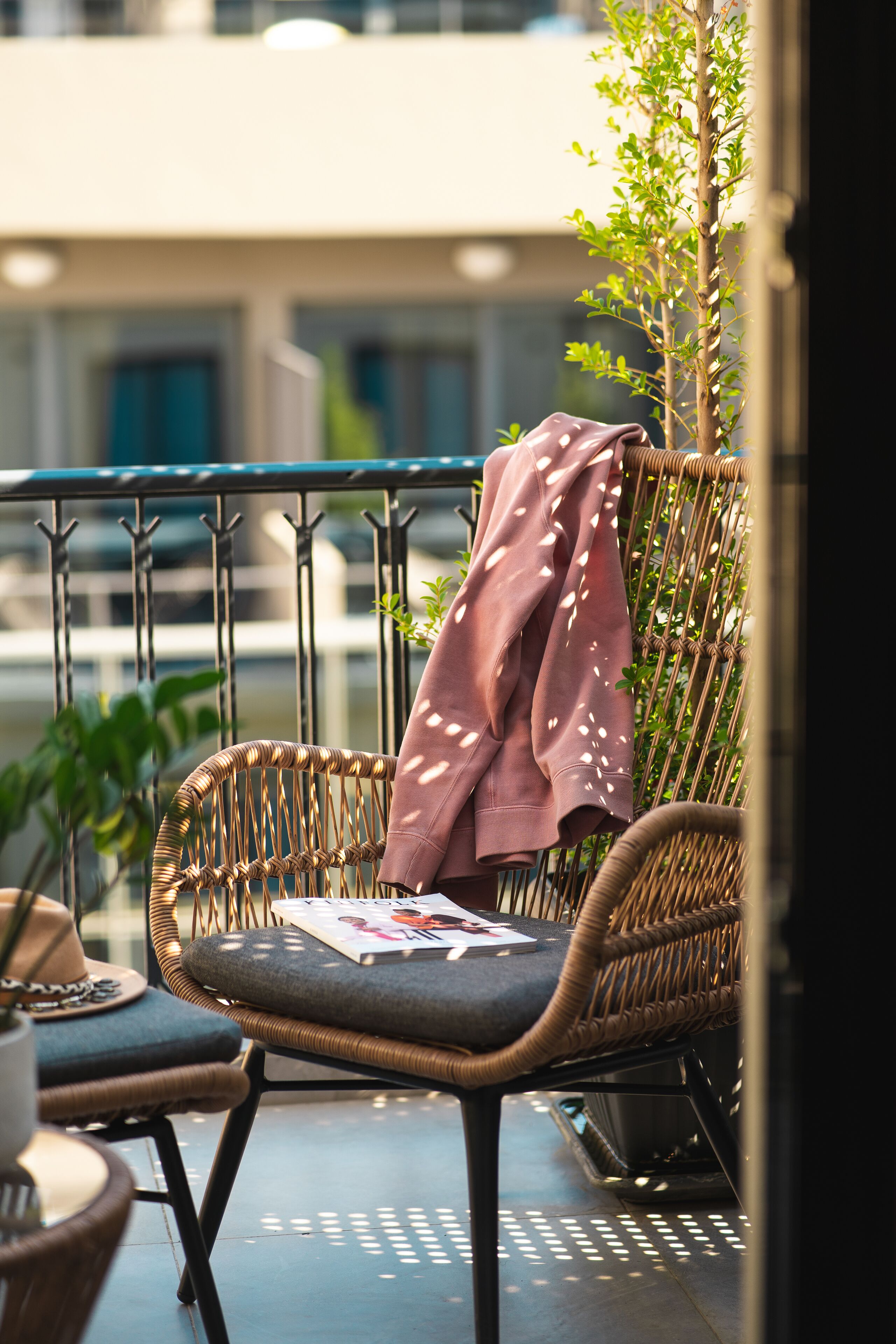 A wicker chair with a cushion and a magazine, a small table with a hat and a cup, and a plant in an urban outdoor setting.