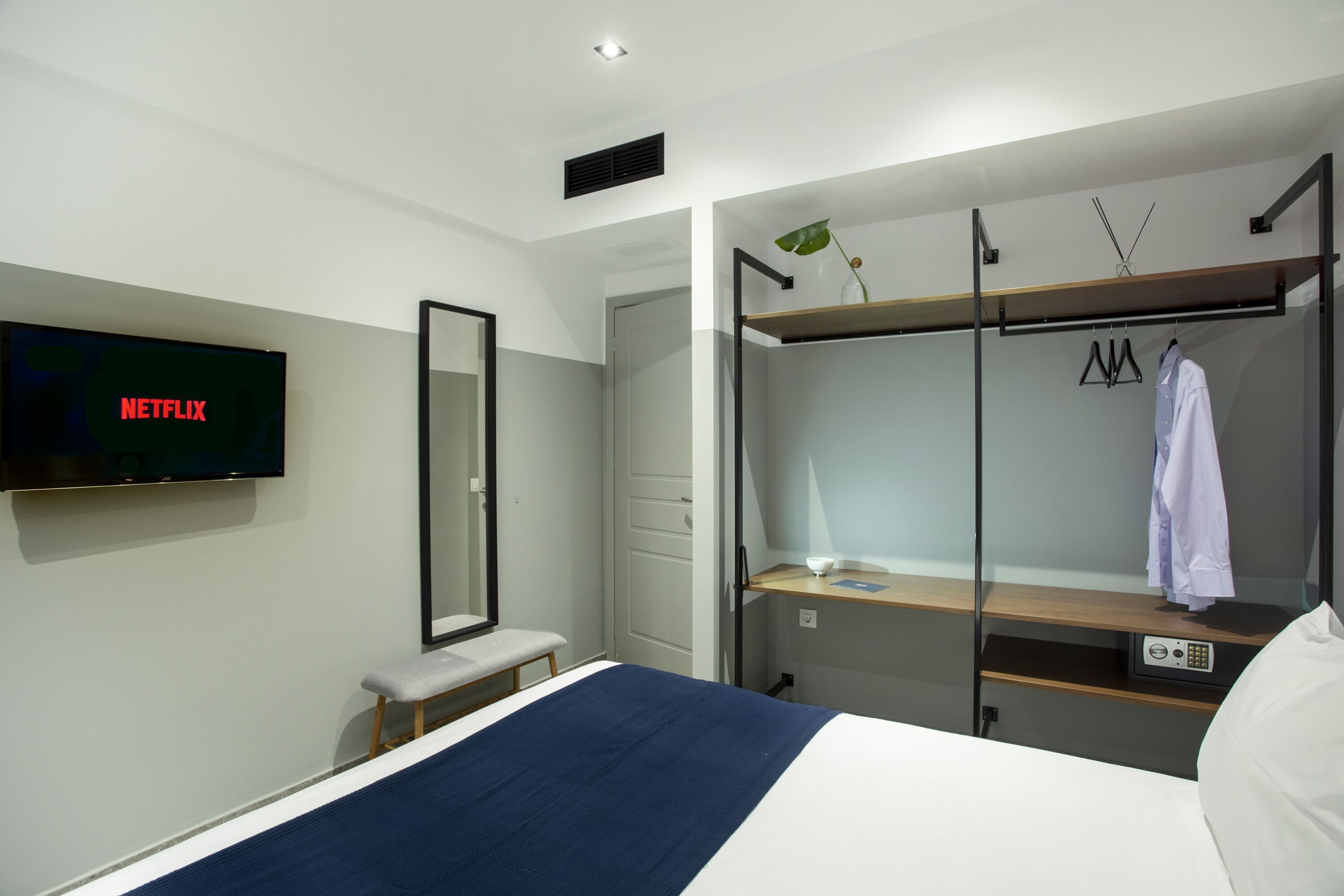 Une chambre d’hôtel comprenant un lit, un téléviseur affichant le logo « NETFLIX », un banc, un miroir, un porte-manteau et un bureau avec une lampe.