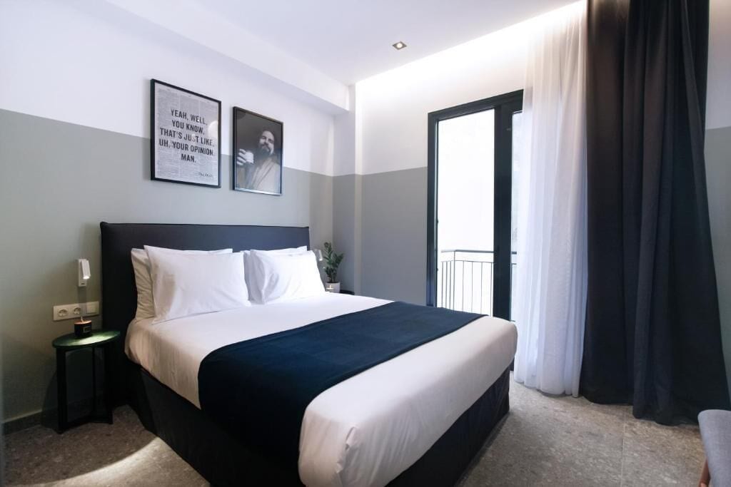 A hotel room with a large bed, a bedside table with a lamp, a framed quote on the wall, and a view of the outdoors through a window.