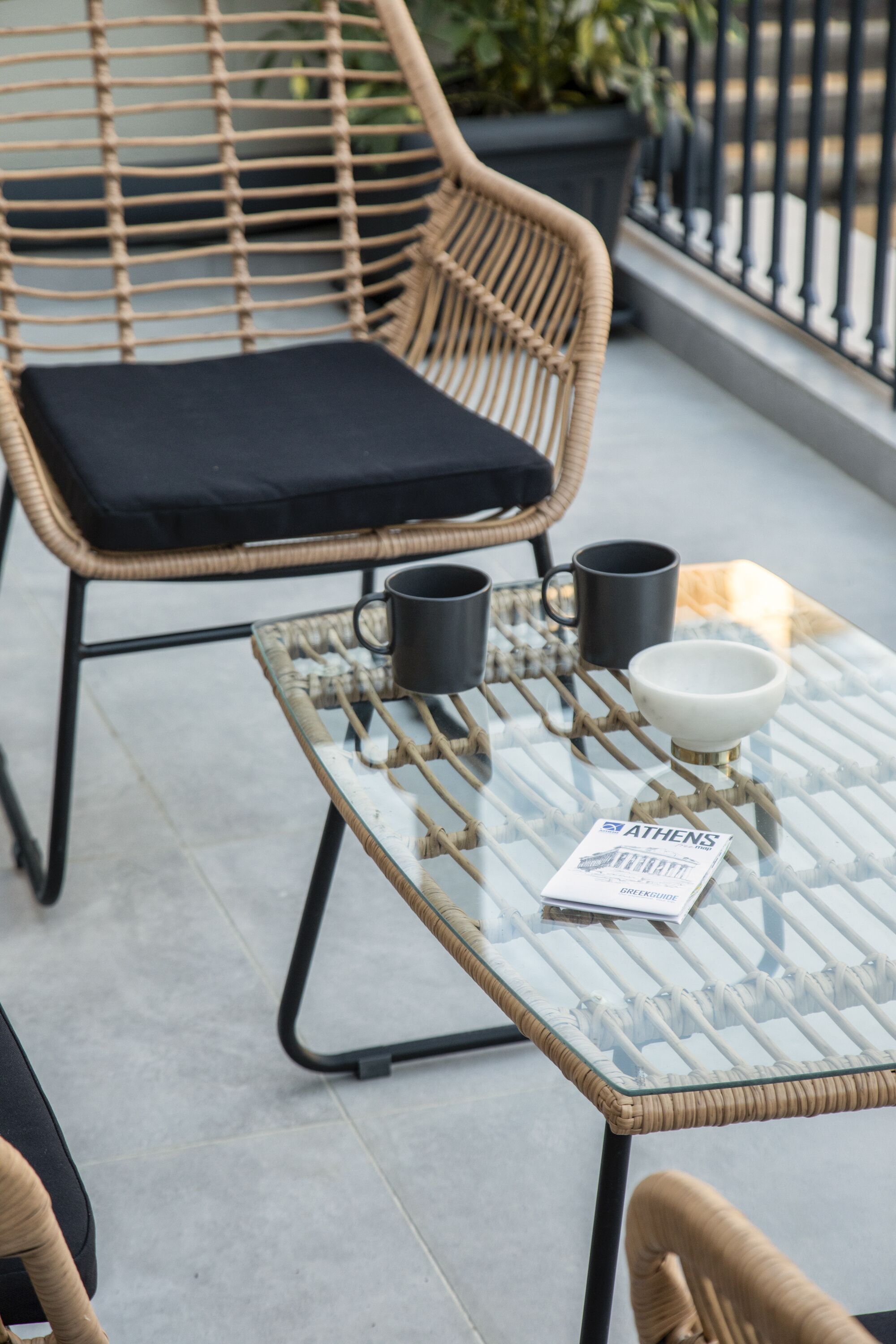 A wicker chair with a black cushion, a glass table with a brochure, and two black mugs.