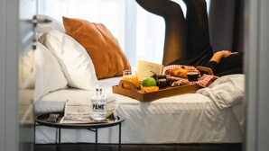 A hotel room with a bed, a bedside table with a newspaper, a tray with food and drinks, and a window with curtains. - Pame Paradiso (Athens)