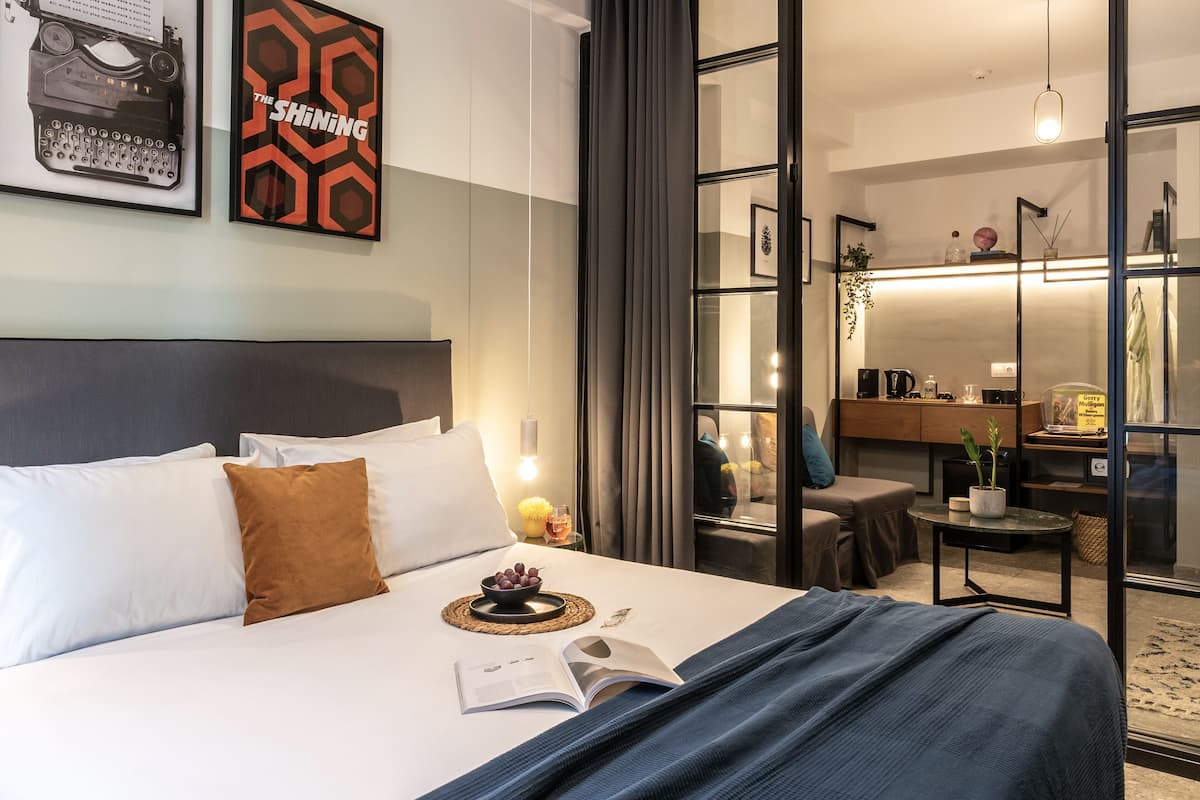 A modern hotel room with a neatly made bed, a bedside table with a lamp, a small side table with a plant, and a view of the city through a large window.