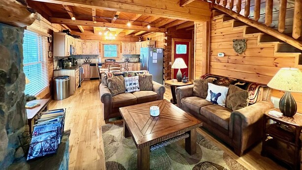 A cabin interior with a wooden staircase, a kitchen area, and a seating area with two sofas and a coffee table. - Bear Necessities - Hot tub, View, Spectacular! (Ferguson)