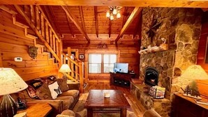 A cozy log cabin interior with a stone fireplace, wooden staircase, and a television in the living area. - Bear Necessities - Hot tub, View, Spectacular! (Ferguson)