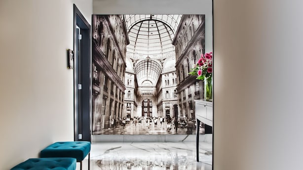 Um corredor com um espelho grande refletindo um amplo espaço arquitetônico, dois bancos acolchoados azuis e um vaso com flores à direita.