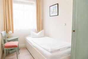 A small, neatly arranged bedroom with a single bed, a chair, a desk, and a window with curtains. - Apartment Rosenhof 01 - Wh. 01 (Kühlungsborn)