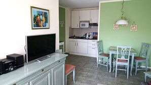 Un espacio habitable compacto con kitchenette, un comedor con mesa y sillas, un televisor en un mueble y un cuadro en la pared.
