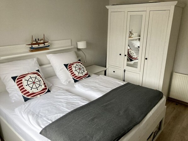 Un lit bien fait, avec une literie blanche et deux coussins décoratifs sur le thème de la marine. Une armoire blanche avec un miroir se trouve à droite du lit.