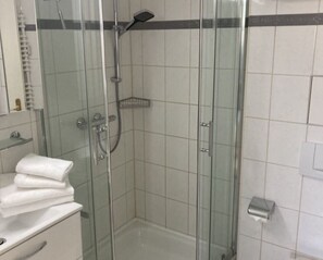 Ducha envuelta en vidrio con cabezal de ducha de mano, puerta de vidrio y pared de azulejos blancos.