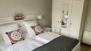 En snyggt bäddad säng med vita sängkläder och två dekorativa kuddar, en vit garderob och en liten modell av ett skepp på nattduksbordet.