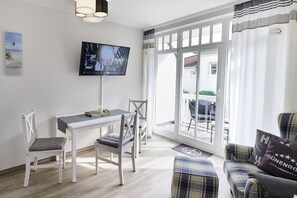 A room with a television mounted on the wall, a dining table with chairs, a sofa, and a large window with curtains. - Rosenhof Apartment 05 DÜNENROSE - Apt. 05 (Kühlungsborn)