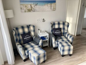 Une pièce comprenant deux fauteuils à motifs écossais et un pouf assorti, une petite table d’appoint blanche et un tableau sur le thème de la plage accroché au mur.