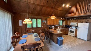 A wooden cabin interior with a dining area, kitchen, and a large window.