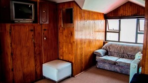 A compact RV interior with a wooden finish, a white bench, a sofa, a television mounted on a cabinet, and a window providing natural light. - Comfit Zone B&B (Newcastle)
