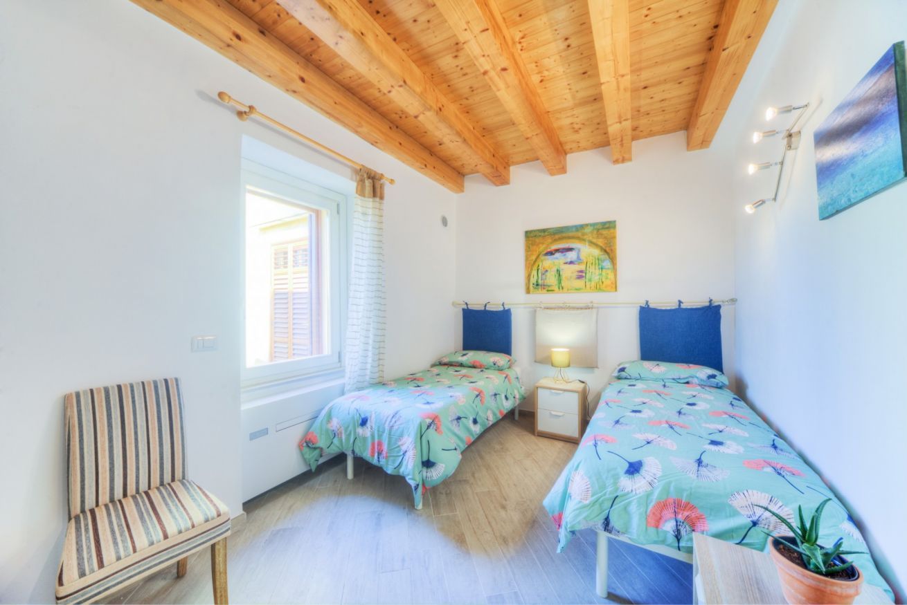 A room with two single beds, a chair, a small table, a window with blinds, a painting on the wall, and a wooden ceiling.