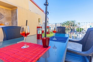 Un balcón con una mesa dispuesta para dos personas, con una copa de vino tinto, una botella de Campari y una botella de Perol.