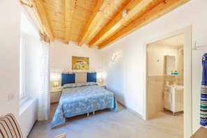 Un dormitorio con techo de madera, una cama con colcha floral, una mesita de noche con lámpara, un cuadro en la pared y un baño con lavabo y espejo.