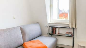 Ein graues Sofa mit einem orangefarbenen Zierkissen, ein schwarzes Bücherregal mit Büchern und ein Fenster mit Vorhängen.