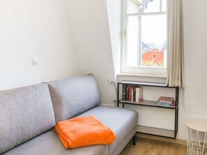 A grey sofa with an orange throw pillow, a black bookshelf with books, and a window with curtains. - Apartment Rosenhof 09 - Wh. 09 (Kühlungsborn)