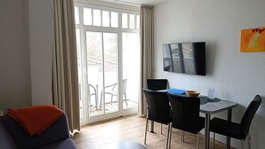 En moderne stue med sofa, lille rundt bord, spiseplads med stole, fjernsyn ophængt på væggen og en skydedør til et udendørs område.