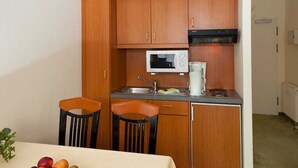 Een compacte keuken met houten kastjes, een magnetron, een koffiezetapparaat en een spoelbak. Er staan twee stoelen bij een tafel met fruit erop.