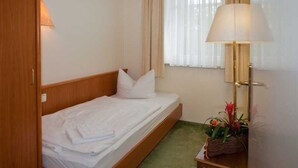 Een enkel bed met een houten hoofdeinde, een bedlampje, een klein tafeltje met een plant en een raam met dunne gordijnen.