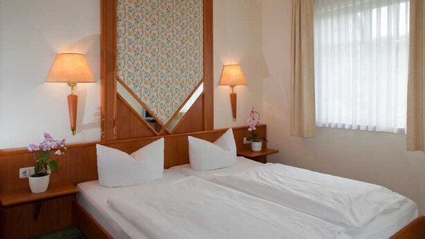 Hotelkamer met een tweepersoonsbed, twee bedlampjes, een spiegel, een potplant en een raam met gordijnen.