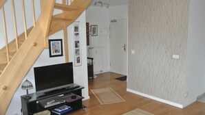 A living room with a wooden staircase, a flat-screen TV on a black stand, a small table with a lamp, and a patterned wall. - Apartment Rosenhof 07 - Wh. 07 (Kühlungsborn)
