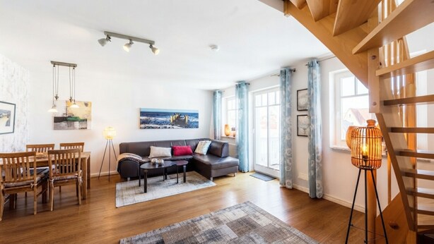 A modern living room with a sofa, dining table, and chairs, a staircase, and a large window with curtains. - Apartment Rosenhof 07 - Wh. 07 (Kühlungsborn)
