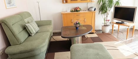 Ein Wohnzimmer mit einem grünen Sofa, einem kleinen Tisch mit einer Vase, einem Holzschrank, einem Fernseher auf einem Schrank und einer Topfpflanze.