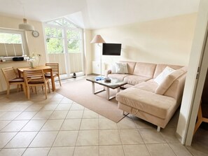 A living room with a beige sofa, a coffee table, a TV mounted on the wall, a dining table with chairs, and a window with blinds.