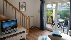 A living room with a TV on a stand, a staircase, a radiator, and a view of the garden through a glass door. - Apartment Rosenhof 12 - Wh. 12 (Kühlungsborn)