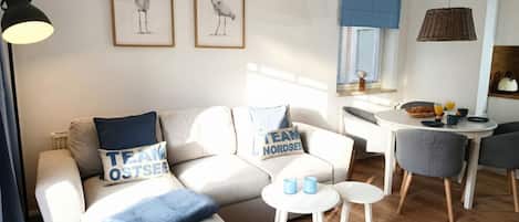 A modern living room with a sofa, a coffee table, and a dining area with chairs. There are framed pictures on the wall and a lamp on the left.