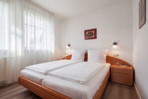 Una cama doble con sábanas blancas, dos mesitas de noche, un aplique en la pared y un cuadro enmarcado.