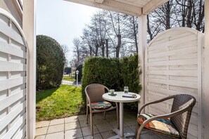 A patio area with a round table and two wicker chairs, a white fence, and a view of a street with trees. - Apartment Rosenhof 11 - Wh. 11 (Kühlungsborn)