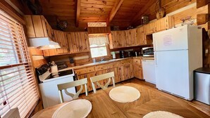 A wooden cabin kitchen with a dining table, wooden cabinets, a refrigerator, and a window with blinds.