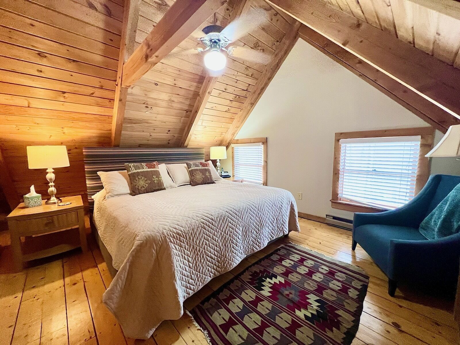 A cozy bedroom with a sloped ceiling, a large bed with patterned bedding, two bedside lamps, a blue armchair, and a patterned rug.