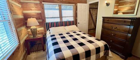Ein Schlafzimmer mit kariertem Bettbezug, Holzwänden, einem Deckenventilator, einem Schminktisch und einem Fenster mit Jalousien.