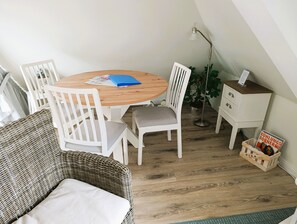 Una habitación acogedora con una mesa redonda de madera, sillas blancas, un sofá de mimbre y un mueble blanco pequeño con una revista.